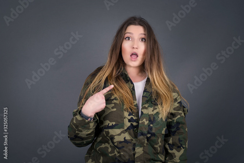 Surprised beautiful female student being in stupor shocked, has astonished expression pointing at herself with finger saying: Who me? Isolated over gray background.