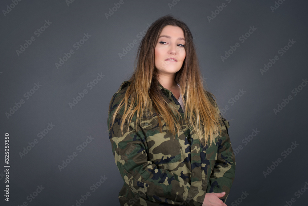 Dreamy young female with thoughtful expression, looks away keeps hands down bitting her lip thinks about something pleasant poses against gray wall. Dreamy young female with thoughtful expression, looks away keeps hands down bitting her lip thinks about something pleasant poses against gray wall.