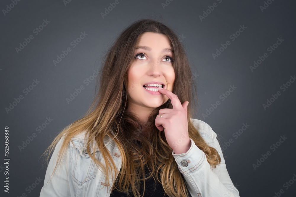 Dreamy female with thoughtful expression, looks to the camera, keeps hand near face, bitting a finger thinks about something pleasant, poses against gray wall. Dreamy female with thoughtful expression, looks to the camera, keeps hand near face, bitting a finger thinks about something pleasant, poses against gray wall.