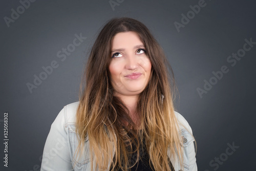 Gloomy, wondering young woman face looking up lips together, being upset thinking about something important, keeps hands down, woman standing outdoors.