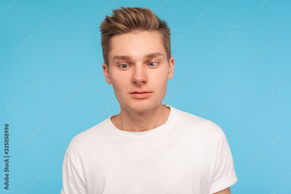 Portrait of funny comic young man in casual white t-shirt looking cross-eyed with awkward silly dumb expression, having fun alone, playing fool. indoor studio shot isolated on blue background, closeup Portrait of funny comic young man in casual white t-shirt looking cross-eyed with awkward silly dumb expression, having fun alone, playing fool. indoor studio shot isolated on blue background, closeup