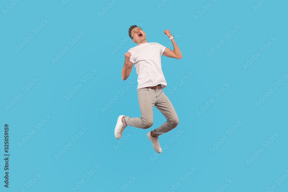 Full length, excited successful man in white t-shirt and casual pants jumping with happiness and shouting yes, I did it, feeling inspired and energetic. indoor studio shot isolated on blue background
