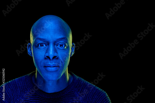 Blue light studio photo of a black man looking at the camera with amazement expression. Isolated on black background. Horizontal with copyspace