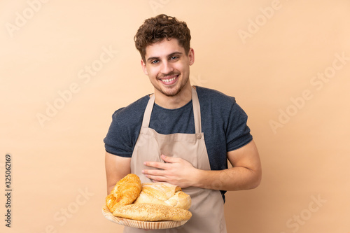 Male baker holding a table with several breads isolated on beige background laughing