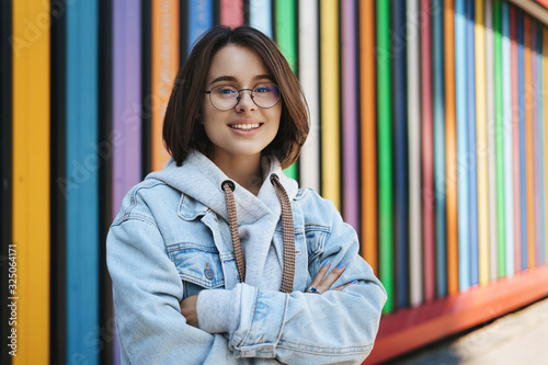 Cheerful young queer girl in glasses, denim jacket, standing near rainbow wall, cross hands over chest and smiling camera happy, lifestyle, urban life and generation concept photo