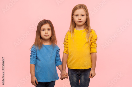 Little friends. Two cute charming preschool girls in sweatshirts standing together, holding hands and looking at camera with serious calm expression. indoor studio shot isolated on pink background