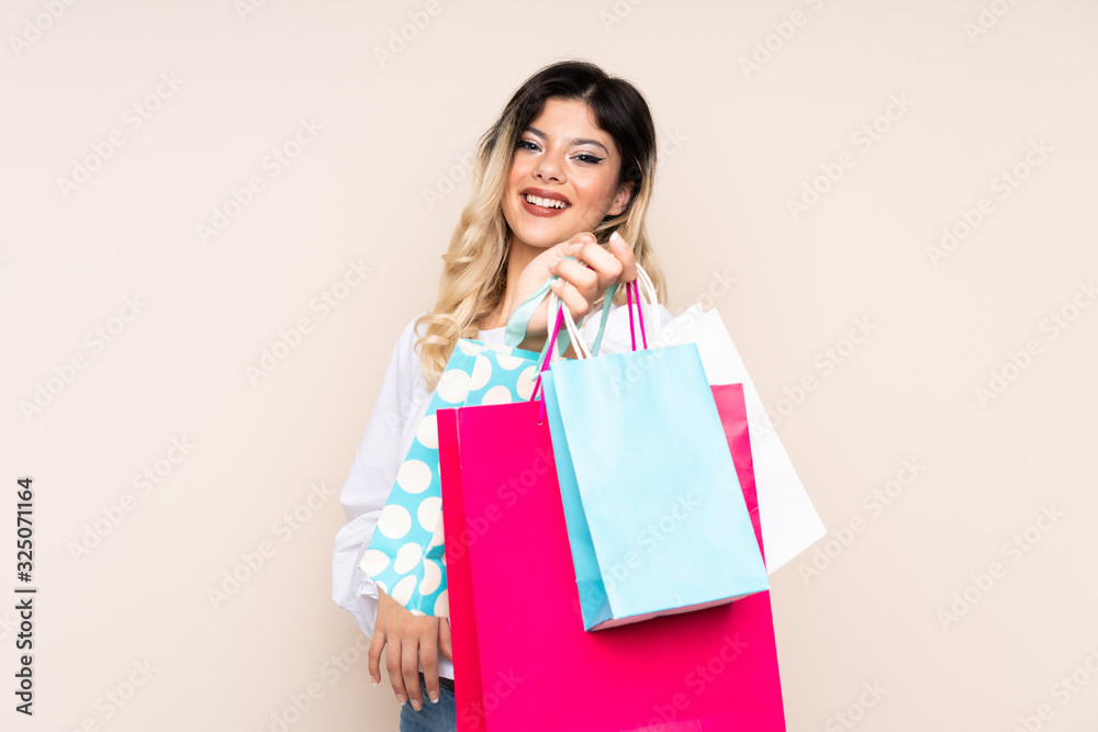 Teenager girl isolated on beige background holding shopping bags and giving them to someone