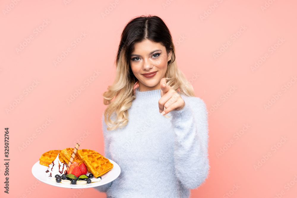 Teenager girl holding waffles on isolated pink background points finger at you with a confident expression Teenager girl holding waffles on isolated pink background points finger at you with a confident expression