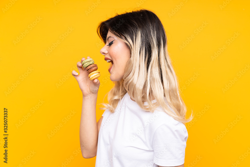 Teenager girl isolated on yellow background holding colorful French macarons and eating it Teenager girl isolated on yellow background holding colorful French macarons and eating it