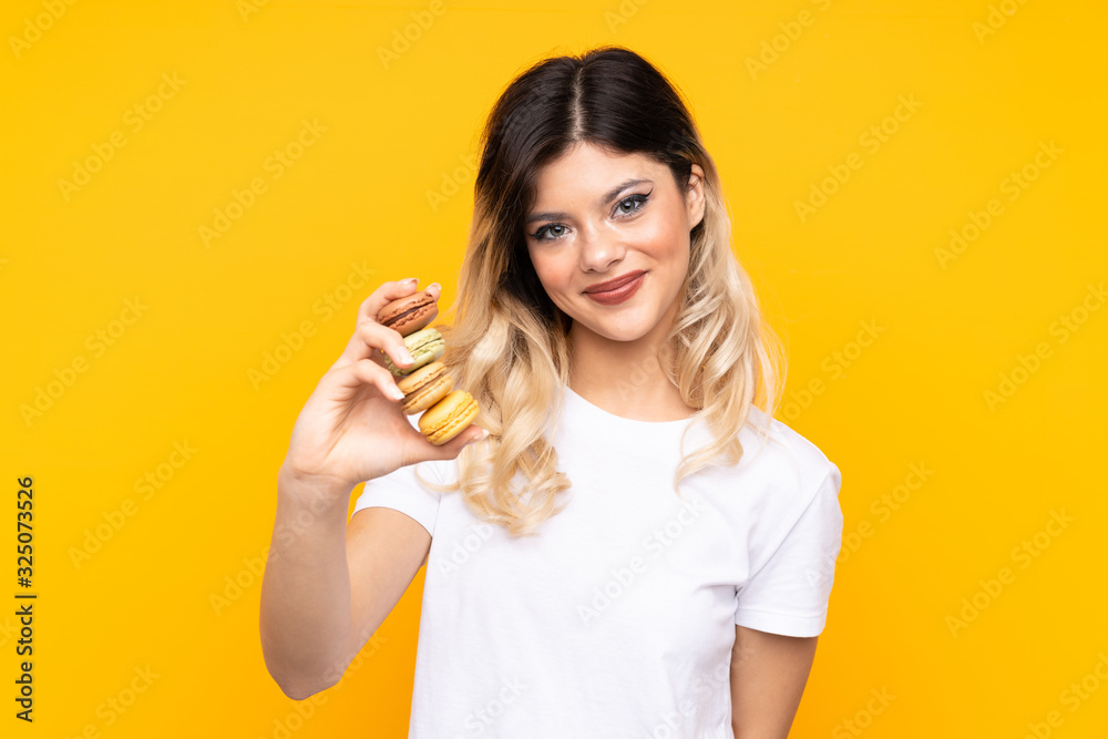 Teenager girl isolated on yellow background holding colorful French macarons and with happy expression Teenager girl isolated on yellow background holding colorful French macarons and with happy expression