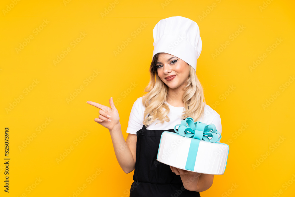 Teenager girl pastry holding a big cake isolated on yellow background pointing finger to the side Teenager girl pastry holding a big cake isolated on yellow background pointing finger to the side