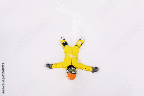Kid snowboarder wearing yellow ski jumpsuit lying on the snow in ski resort