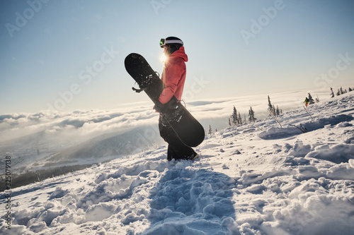 Female snowboarder holding snowboard standing on mountain slop, preparing to snowboarding. Sunny winter day in ski resort