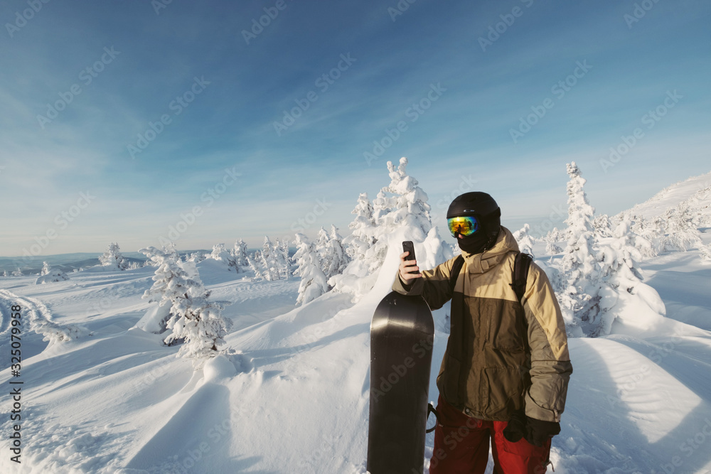 Snowboarder using  mobile phone in the mountains in winter. Beautiful snow covered fir forest on background