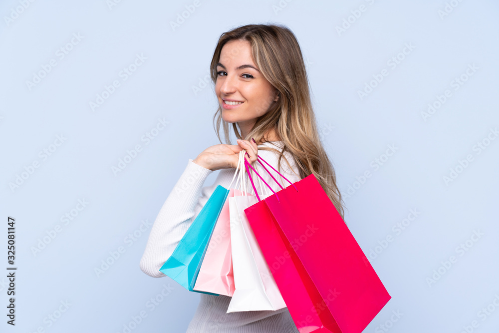 Young woman over isolated blue background holding shopping bags and smiling
