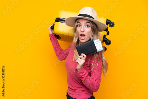 Young woman over isolated yellow background in vacation with suitcase and passport and surprised