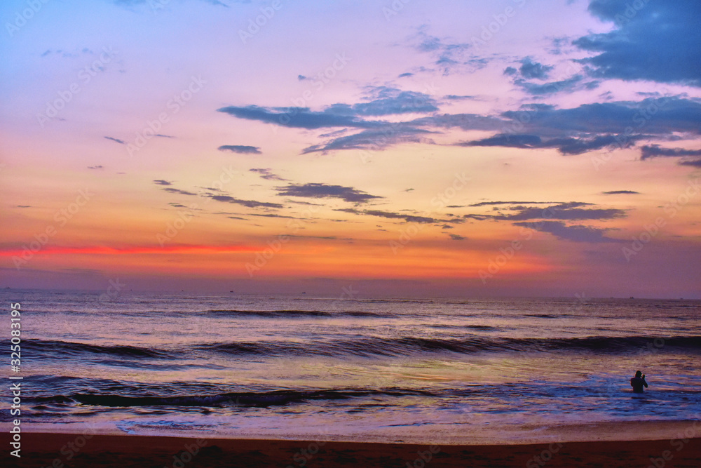 a man in the water under the colorful sky with small waves in a beach in goa