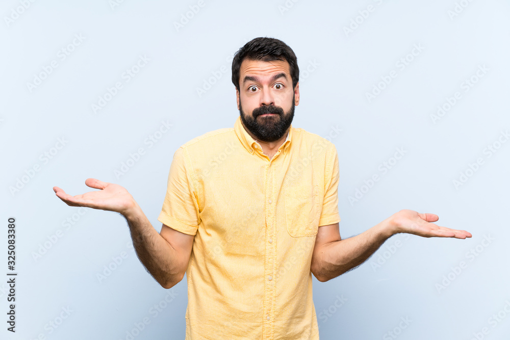 Young man with beard over isolated blue background having doubts while raising hands Young man with beard over isolated blue background having doubts while raising hands