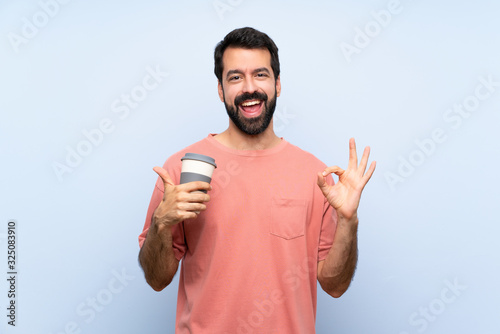 Young man with beard holding a take away coffee over isolated blue background showing ok sign and thumb up gesture