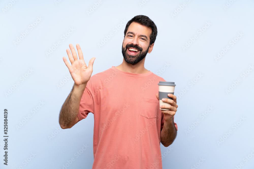 Young man with beard holding a take away coffee over isolated blue background saluting with hand with happy expression