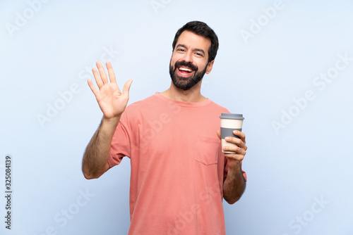 Young man with beard holding a take away coffee over isolated blue background saluting with hand with happy expression