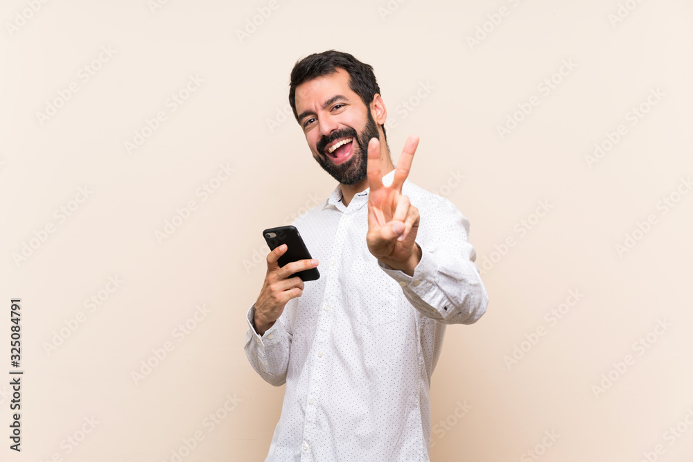 Young man with beard holding a mobile smiling and showing victory sign Young man with beard holding a mobile smiling and showing victory sign