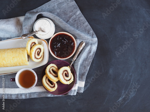 Homemade biscuit sponge roll with sweet plum jam and tea against a dark background. Natural homemade dessert. Top view, place for text.