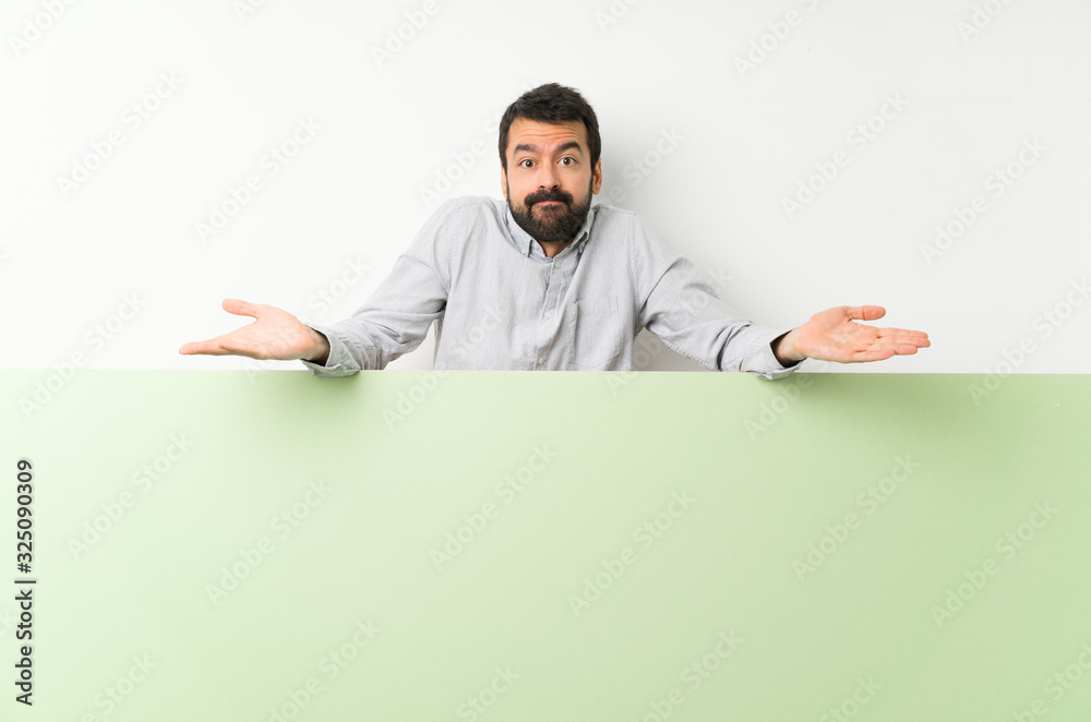 Young handsome man with beard holding a big green empty placard having doubts while raising hands Young handsome man with beard holding a big green empty placard having doubts while raising hands