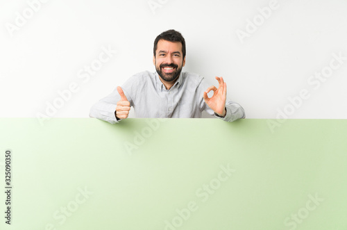 Young handsome man with beard holding a big green empty placard showing ok sign and thumb up gesture