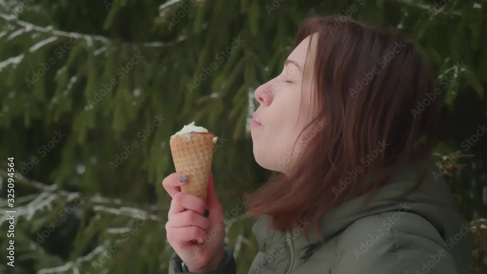 view of woman in green coat eating ice-cream with pleasure. She smiles and enjoys dessert. Summer tastes in cold winter weather. Green fur tree on background. view of woman in green coat eating ice-cream with pleasure. She smiles and enjoys dessert. Summer tastes in cold winter weather. Green fur tree on background.