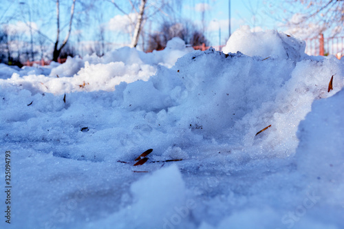 Dirty snow in a closeup during springtime. In this photo you can see melting snow, some small rocks, plants pieces and other debri. Trees and sun in the background