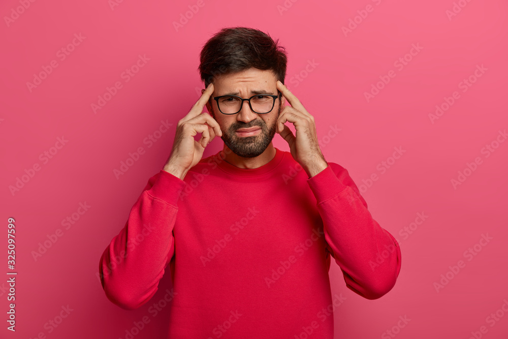 Troubled distressed bearded man keeps index fingers on temples to focuse, tries to concentrate on task, frowns and grimaces, has huge headache or migraine, bothered with painful disease, models indoor Troubled distressed bearded man keeps index fingers on temples to focuse, tries to concentrate on task, frowns and grimaces, has huge headache or migraine, bothered with painful disease, models indoor
