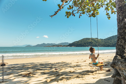 Beautiful girl swinging on wooden swing on the tree at the beach, happy smiling girl with red hair, playing on the beach on vacation trip, at Parnaioca beach, in Ilha Grande, in Rio de Janeiro, Brazil photo