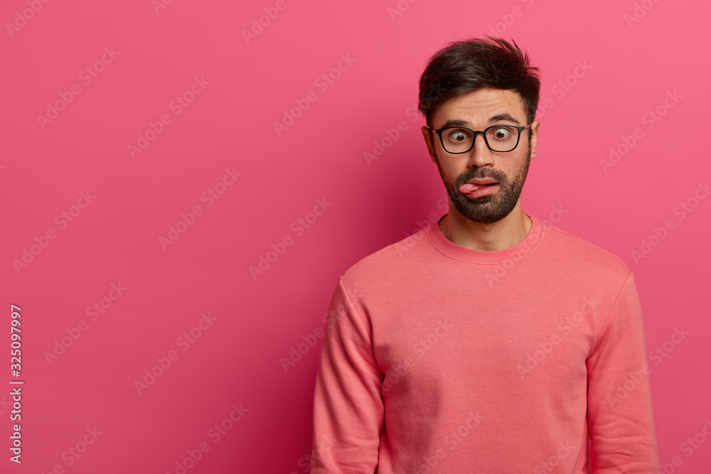 Crazy funny bearded man crosses eyes and sticks out tongue, has bored expression, foolishes around, wears spectacles and casual jumper, isolated on pink background. Postive human feelings concept Crazy funny bearded man crosses eyes and sticks out tongue, has bored expression, foolishes around, wears spectacles and casual jumper, isolated on pink background. Postive human feelings concept