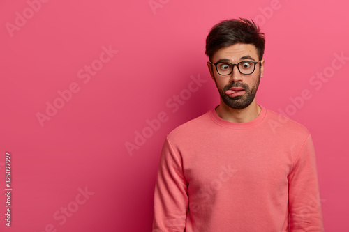 Crazy funny bearded man crosses eyes and sticks out tongue, has bored expression, foolishes around, wears spectacles and casual jumper, isolated on pink background. Postive human feelings concept