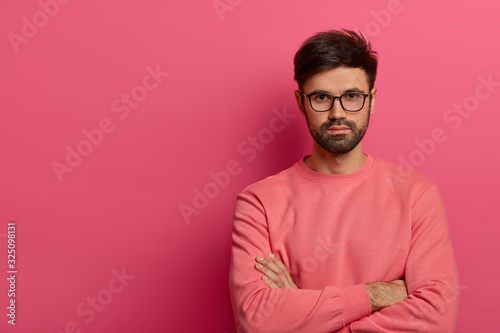 Self confident serious bearded man stands with arms folded, looks seriously at camera, going to discuss interesting topic with interlocutor, wears optical glasses and sweater, isolated on pink wall