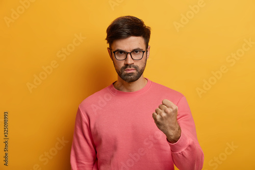 Serious irritated aggressive man holds fist up, expresses negative feelings and attitude, threatening someone, wears spectacles and sweater, isolated over yellow background. I will show you gesture