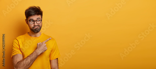 Doubtful man points at upper right corner, feels hesitant about something, purses lips with confusement, poses against yellow backgroud, wears casual clothes. Advertising and people concept.