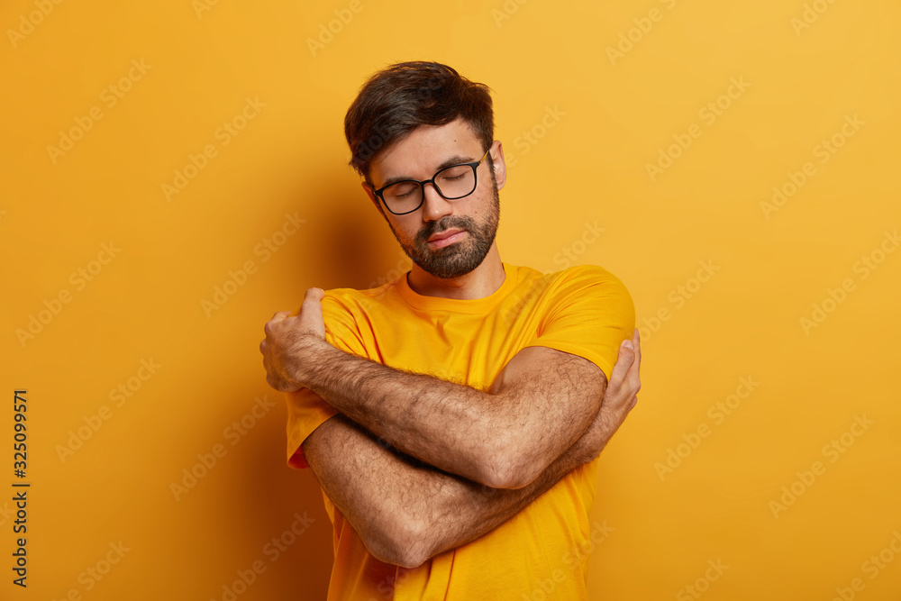 Relaxed unshaven man embraces himself, keeps arms around shoulders, expresses self love, wears casual clothes, recalls something pleasant, isolated on yellow background, feels comfort, home tenderness Relaxed unshaven man embraces himself, keeps arms around shoulders, expresses self love, wears casual clothes, recalls something pleasant, isolated on yellow background, feels comfort, home tenderness