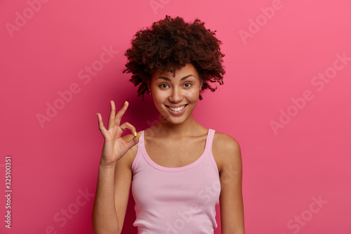 Pretty dark skinned female model shows okay gesture, reassures everything goes excellent, demonstrates no problem sign, gives positive feedback or likes idea, wears casual tank top, isolated on pink