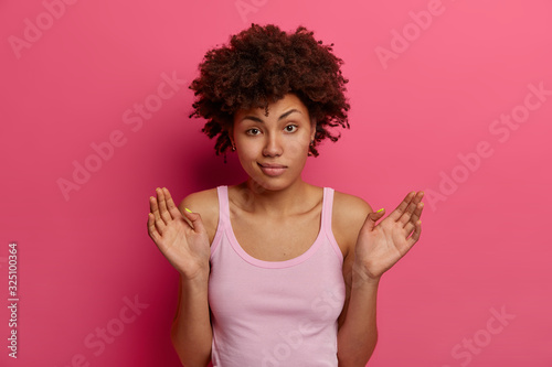 Indifferent hesitant woman with natural curly hiar, raises plams and feels apathy, wears casual tank top, looks confusingly at camera, poses over pink vibrant wall, has no explanation to what happened