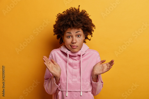 Indecisive curly woman raises palms in clueless gesture, looks doubtfully, faces difficult choice, wears casual anorak, has doubts, considers if she did right, has questioned expression, poses indoor