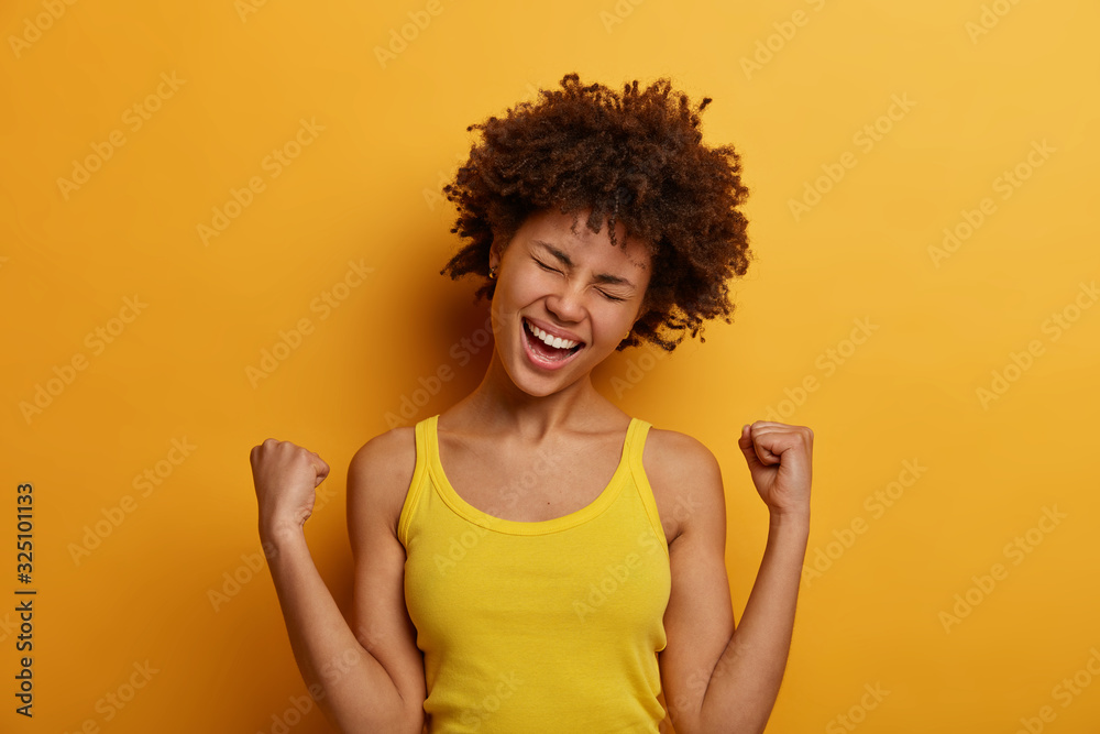Triumphing woman rejoices winning contest, tilts head and laughs out positively, wears casual tank top, clenches fists, celebrates victory, finally achieves goal, isolated over yellow background Triumphing woman rejoices winning contest, tilts head and laughs out positively, wears casual tank top, clenches fists, celebrates victory, finally achieves goal, isolated over yellow background