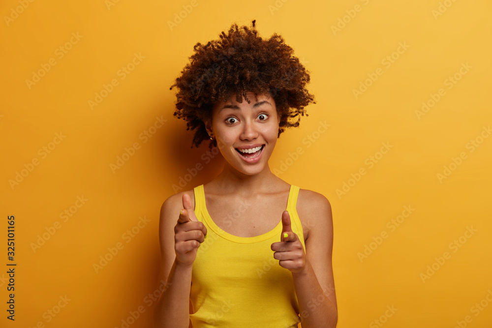 Check it out! Portrait of joyful excited young African American woman points index fingers directly at camera, says great offer for you, shows gun sign dressed casually isolated over yellow background Check it out! Portrait of joyful excited young African American woman points index fingers directly at camera, says great offer for you, shows gun sign dressed casually isolated over yellow background