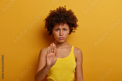 Beautiful serious young woman pulls hand towards camera in stop gesture  asks to calm down and stop worrying  demands to hold and makes prohibition  wears casual clothes  yellow color prevails