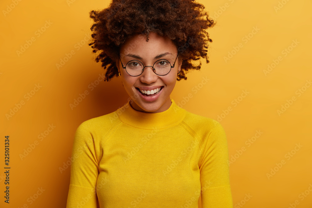 Cropped image of beautiful glad dark skinned model smiles broadly, being in high spirit, enjoys free time, spends weekend with boyfriend, wears transparent glasses and sweater, isolated on yellow wall Cropped image of beautiful glad dark skinned model smiles broadly, being in high spirit, enjoys free time, spends weekend with boyfriend, wears transparent glasses and sweater, isolated on yellow wall