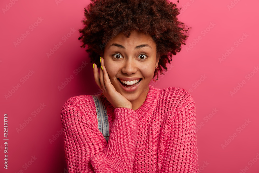 Close up shot of positive young Afro American woman keeps hand on cheek, smiles broadly, enjoys good time, has happy expression, shows white teeth, wears knitted sweater, isolated on pink wall Close up shot of positive young Afro American woman keeps hand on cheek, smiles broadly, enjoys good time, has happy expression, shows white teeth, wears knitted sweater, isolated on pink wall