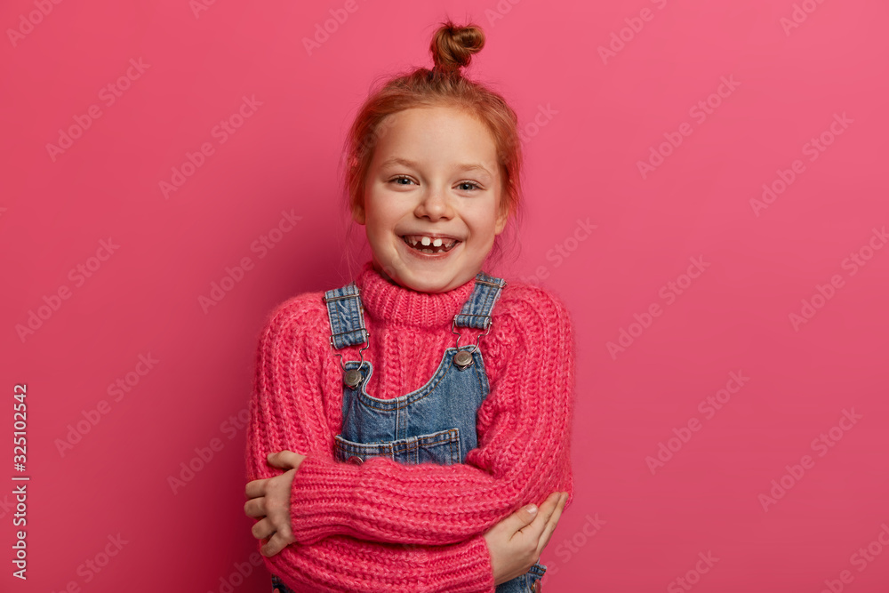 Cheerful small ginger girl hugs herself, feels comfortable, has new woolen pink sweater, warm soft outfit, smiles toothily, shows missing teeth, has ginger hair, isolated on pink background. Cheerful small ginger girl hugs herself, feels comfortable, has new woolen pink sweater, warm soft outfit, smiles toothily, shows missing teeth, has ginger hair, isolated on pink background.
