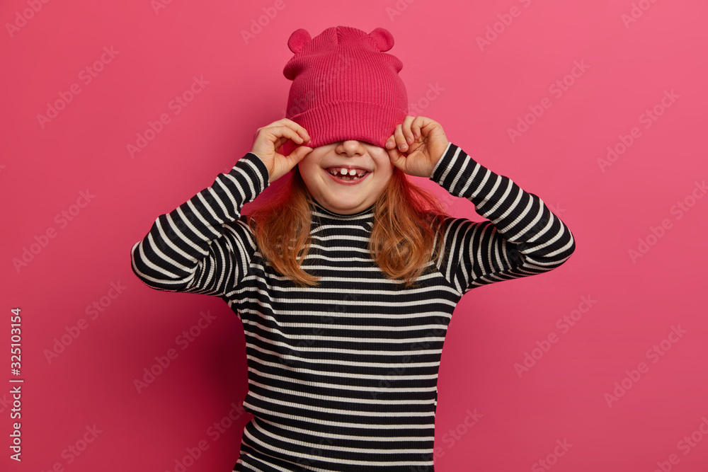 Isolated shot of pretty four years old girl wears striped jumper and pink hat, has fun and covers eyes, enjoys spending time in family circle, isolated on pink background. Children, emotions, clothes Isolated shot of pretty four years old girl wears striped jumper and pink hat, has fun and covers eyes, enjoys spending time in family circle, isolated on pink background. Children, emotions, clothes