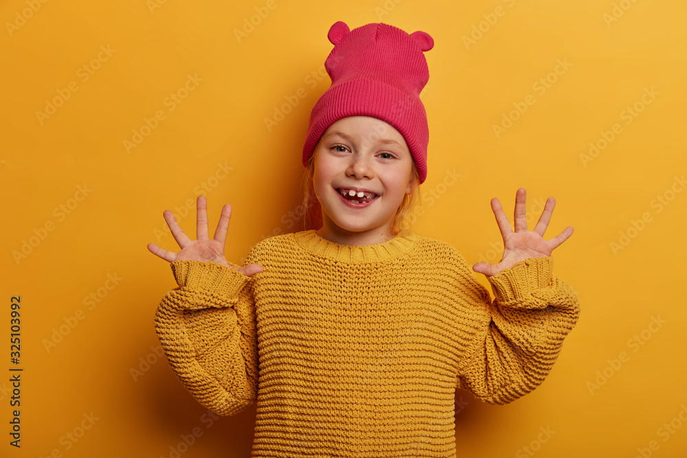 Be positive and keep smiling. Glad adorable European child raises hands and shows palms, expresses good emotions, plays with someone, dressed in knitted sweater, isolated on yellow background